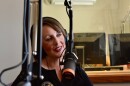 A woman with short brown hair and headphones on sits in a recording studio in front of black computer monitors talking into a hanging microphone.