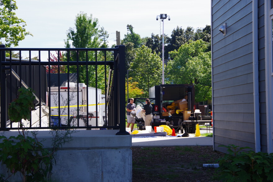 Two individuals can be seen holding a large, plastic object near several trailers in a building parking lot.