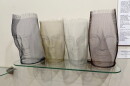 Aluminum wire sculptures of four different heads on a glass shelf and in front of a white wall.