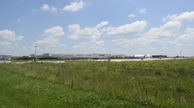 A large warehouse like facility sits behind fields of wild grass and weeds.