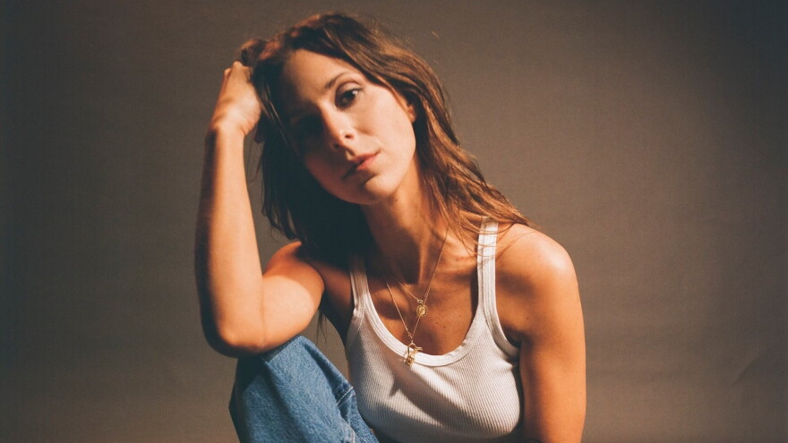 A woman in a white tank top and blue jeans looks at the camera with her elbow resting on her knee and her hand holding her head.