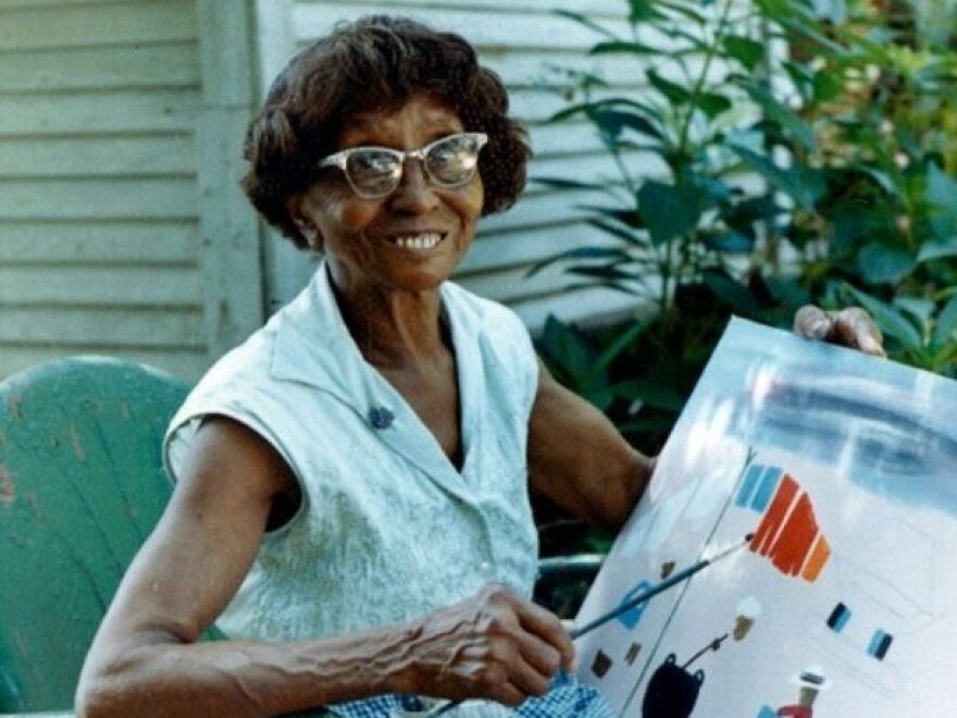 Clementine Hunter holding her artwork. Hunter worked as a house worker at Melrose Plantation, where she started out quilting and doing other crafts before an artist visiting the property gave her leftover paint and brushes. Body - 4