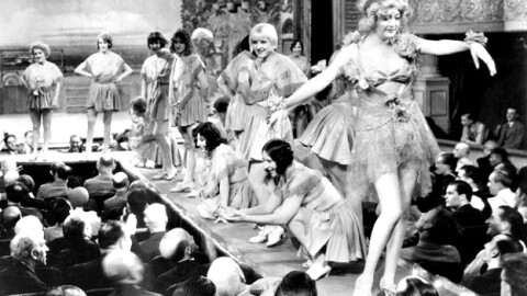 A black and white photograph showing a scene from the 1929 movie Applause directed by Rouben Mamoulian, featuring several showgirls performing on a raised stage before a seated theater audience.
