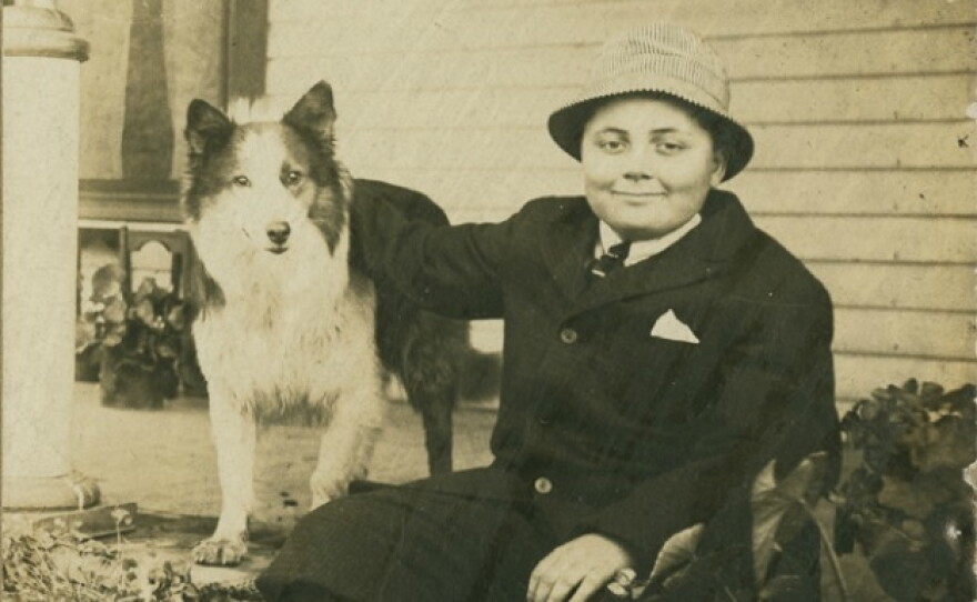 A monochromatic photograph of a young Herman B Wells wearing a dark suit jacket and a brimmed hat, seated outdoors while resting his arm on his standing, long-haired collie dog.