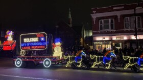 A lighted Christmas float with Snoopy, a Carbondale logo, Snowman and horses leading the float pictured at night during a parade.