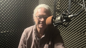 A man with long grey hair tied into a ponytail  and glasses sits behind the microphone in a studio with padded walls.