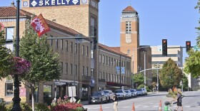 Exterior photo of a city street shows two people crossing the street. Buildings can be seen in the background with cars parallel parked in front of them.
