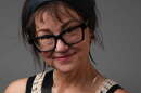 A woman with short brown hair and chunky black glasses smiles at the camera in front of a grey background