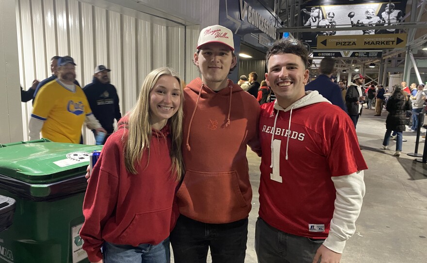 Three young adults, two men and one woman, smile and pose together indoors at a stadium. All are wearing red tops, with the man on the right in a "Redbirds" jersey. Other people and concession stands are visible in the background.