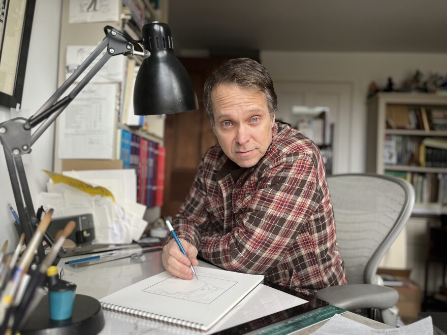 A man with short salt and pepper hair is wearing a plaid button-down shirt and drawing on a pad while sitting at a desk in an office.
