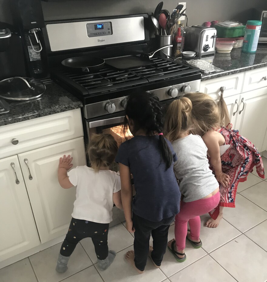 Four small children stand side by side peering into an oven in a kitchen.