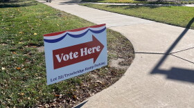 A red, white and blue Vote Here sign beside a campus sidewalk, with trees and a cyclist in the background.