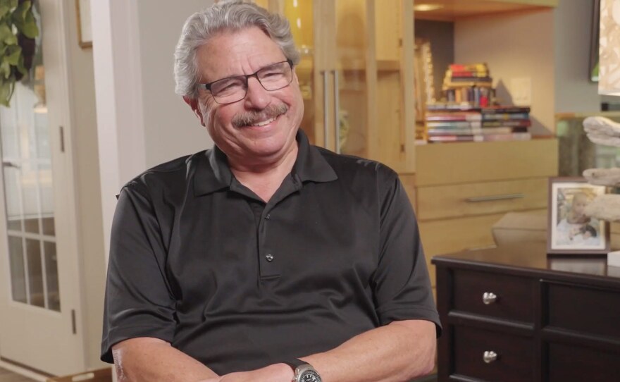 A smiling man with short grey hair and a grey mustache is wearing eyeglasses and a dark grey polo shirt and sitting in a living room.