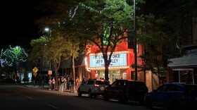A theater with a bright red marquee glows brightly at night framed by dark trees and a night sky. People stand under the theater sign. 