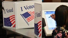 A person uses a stylus to mark a touchscreen ballot. They are sitting inside a booth with a sign that reads "I voted" on it.