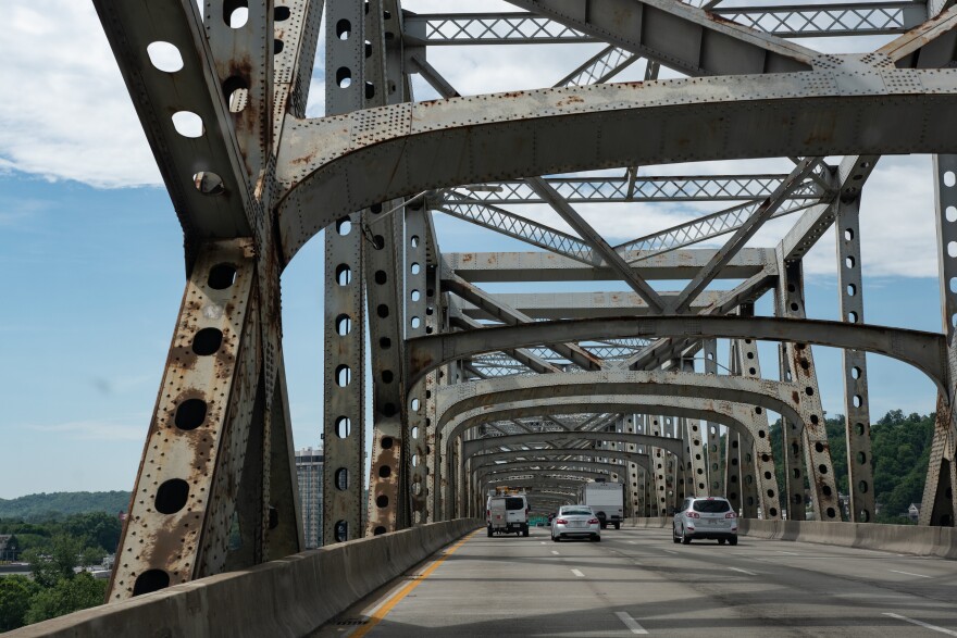 A picture of a bridge taken from the bridge itself. Metal trusses arch above the roadway and cars drive away from the camera in multiple lanes of traffic.