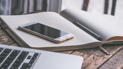 An iPhone sits on a notebook next to a laptop computer.