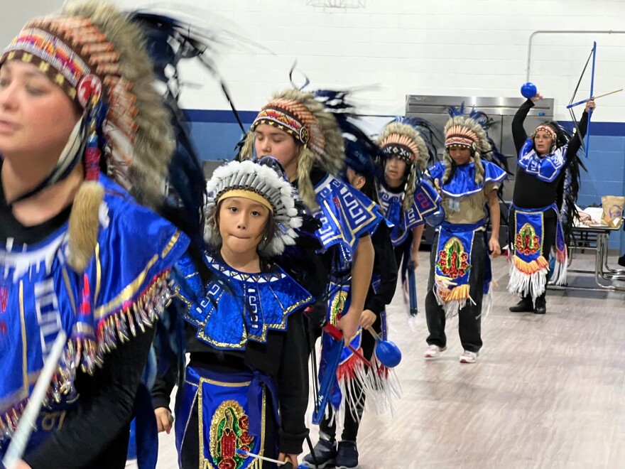 A line of dancers in blue outfits and feathered headdresses prepare for their next dance.