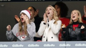 Three women in foreground are wearing cold weather clothes and cheering from inside football stadium club seating. The woman on far left (Taylor Swift) is waring a knitted white and red stocking cap.