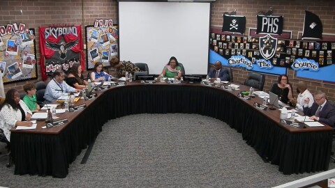 A group of people sits around a large U-shaped table in a meeting room with school-themed decorations and a blank projector screen on the back wall.