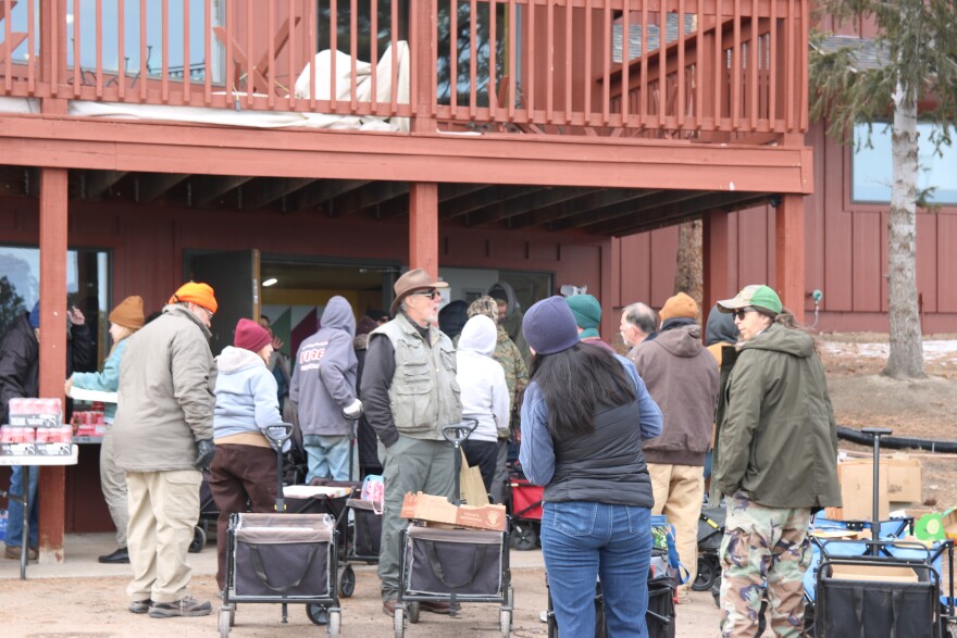 People in winter coats and sweatshirts stand in a line, some carrying the handles of canvas-sided wagons, outside a big red church building.