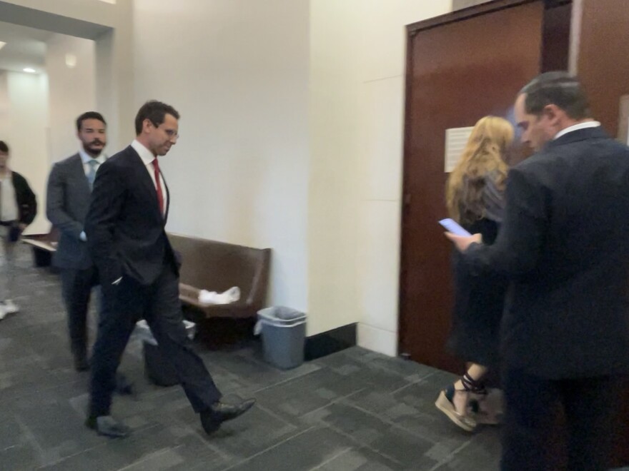 James Fishback, wearing a suit, walks down a courthouse hallway toward a courtroom door, accompanied by others in business attire.