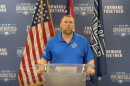 A man wearing a blue shirt stands at a glass podium with flags behind him.