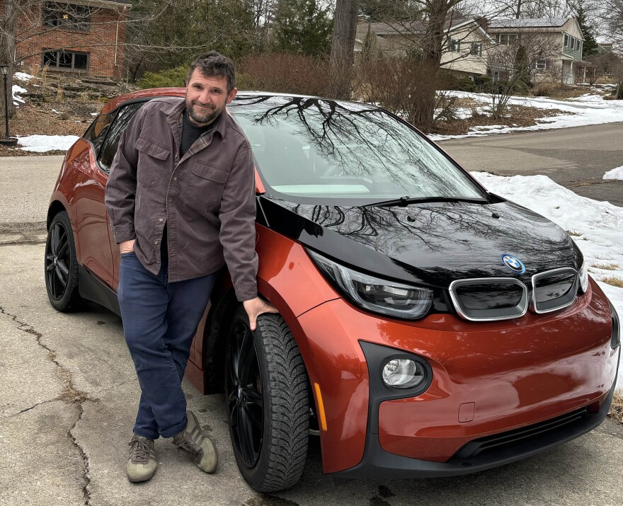 Tyler Lake in a brown chore shirt and jeans standing next to a reddish-brown and black BMW car in a concrete driveway. One hand is in his pocket, one is grabbing a front tire. View of a suburban neighborhood with lingering snow is visible in the background. 