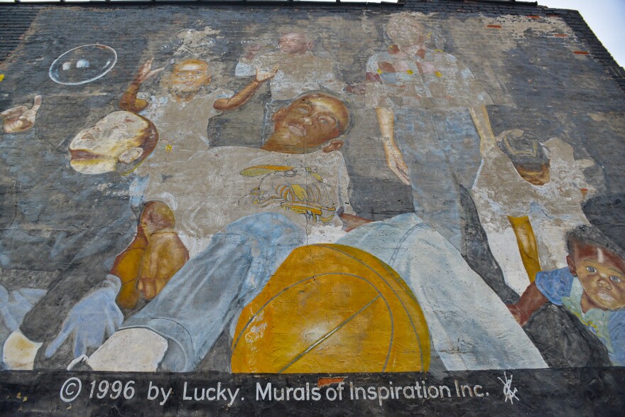 Detail of a faded mural on a black, brick wall shows children of different ages and genders playing. One young boy sits straddling a basketball, another boy is about to pop a bubble, a young girl reaches to clasp other bubbles.