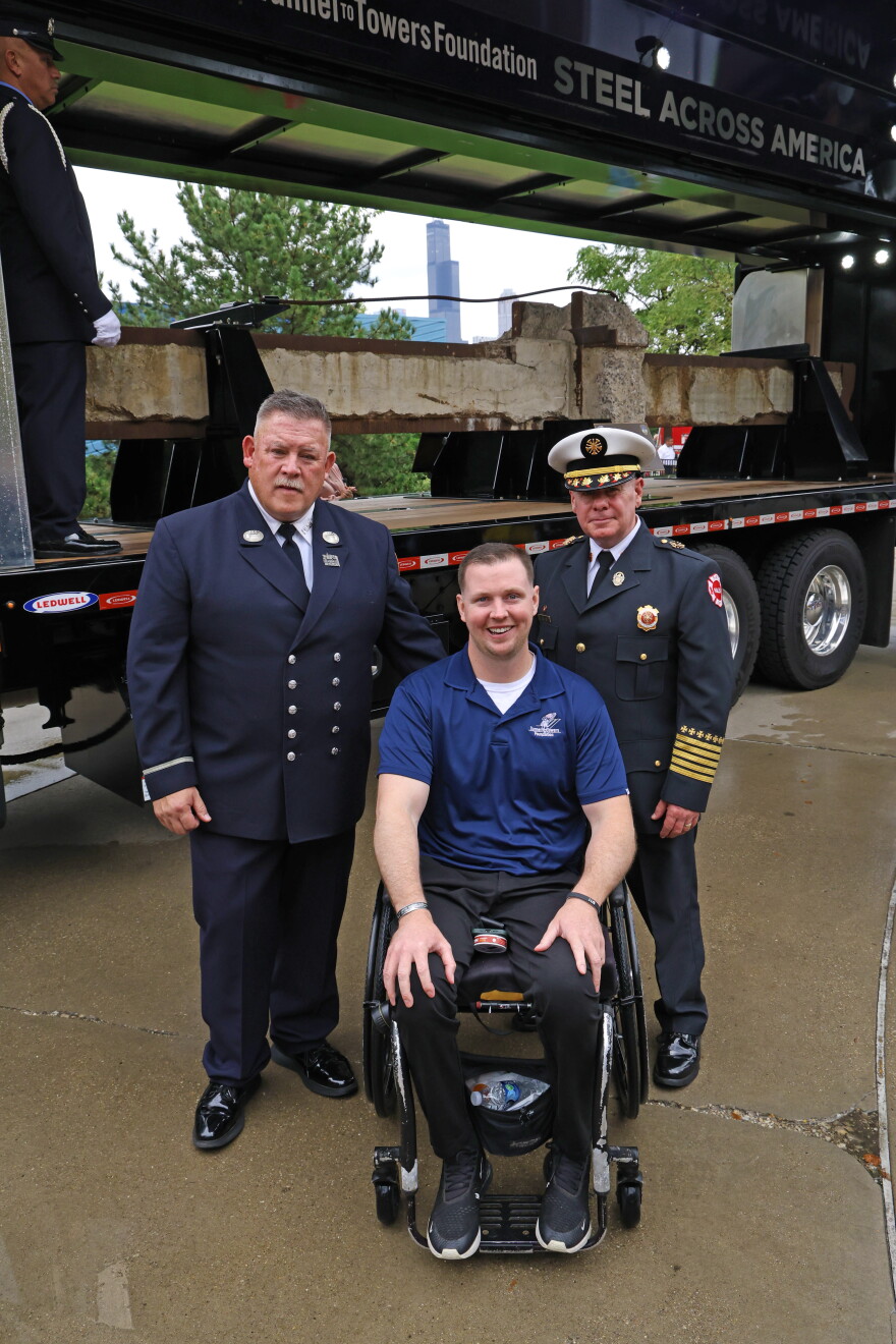 Two FDNY members and a man in a wheelchair pose for a picture by a steel beam from Ground Zero