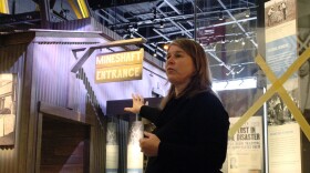 Molly Kruckenberg, executive director of the Montana Historical Society, gestures while leading a tour through the Montana Heritage Center. She stands near a life-size exhibit entrance labeled “Mineshaft Entrance,” with interpretive panels and historical images visible in the background.