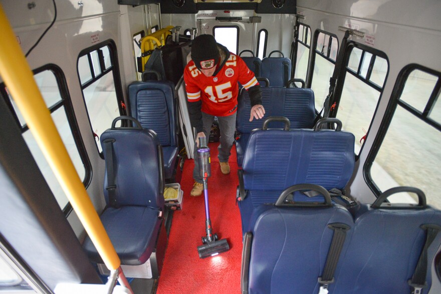 A man wearing a read football jersey with the number 15 printed on it vacuums inside a small bus. The seats are blue and the carpet is red.