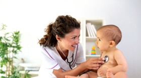 Physician examining her baby patient with stethoscope as the baby sits upright during medical examination.