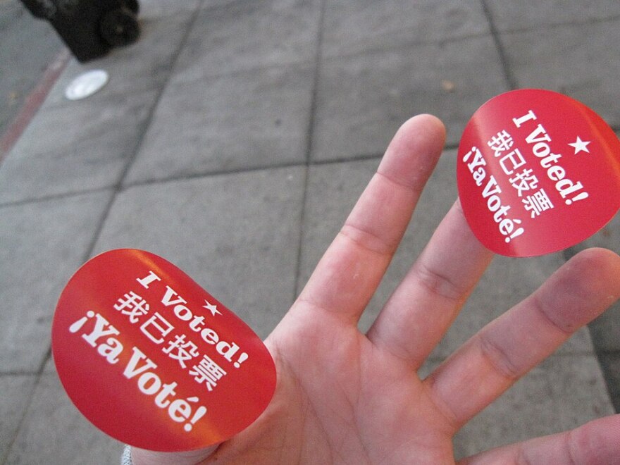 A photo of a hand with light skin holding two stickers from San Francisco that are red with white text. They say "I voted" in English, Chinese and Spanish. 
