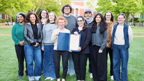 A group of students pose with their instructor in the left corner and two in the center holding a document. They are standing on a grassy lawn with bright daytime light and a brick university building with trees and red brick walkways in the background.