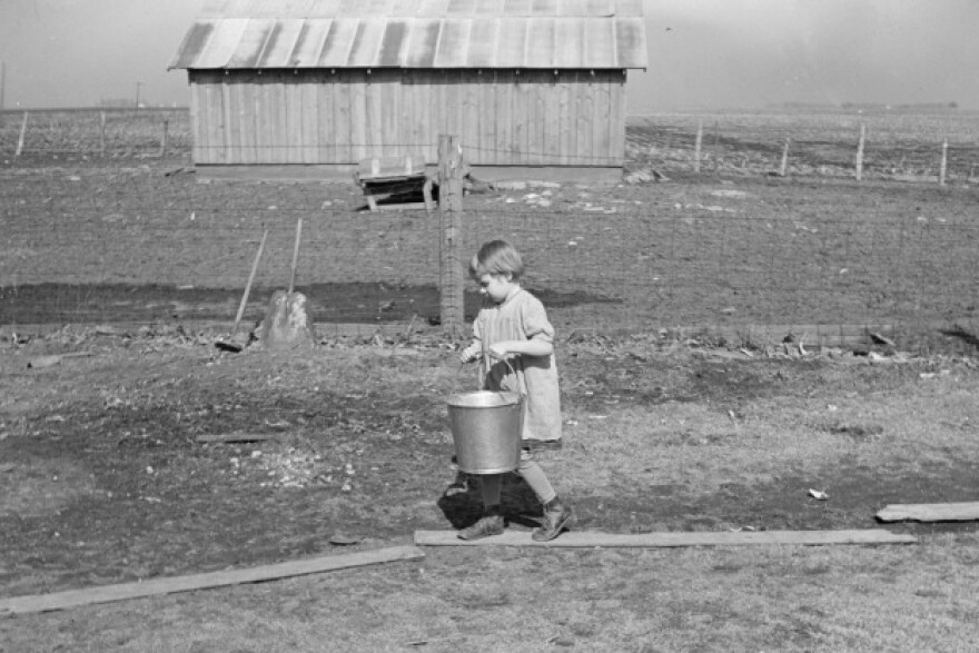A black and white photograph shows a young child walking on a path of wooden boards while carrying a metal bucket. In the background, there is a simple wooden shed or barn, and a wire fence separates the foreground from a flat, open field.