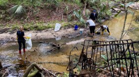 Four people stand in a creek with bags and tools for cleaning up.