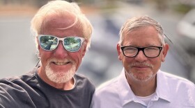 Rich Guy and James Oscarson pose outside James' Tesla in the St. George Sunshine. Each looks a little windblown.  The background is out of focus. Rich stands holding the phone, which can be seen reflected in the white sunglasses he wars. Rich has on a dark grey long-sleeved tee shirt.  He has short white hair and a white goatee.  James is slightly shorter than Rich and wears black framed glassses.  His hair is salt and pepper gray, and he also sports a goatee. James wears a lilac checked button-up shirt with a white tee underneath.