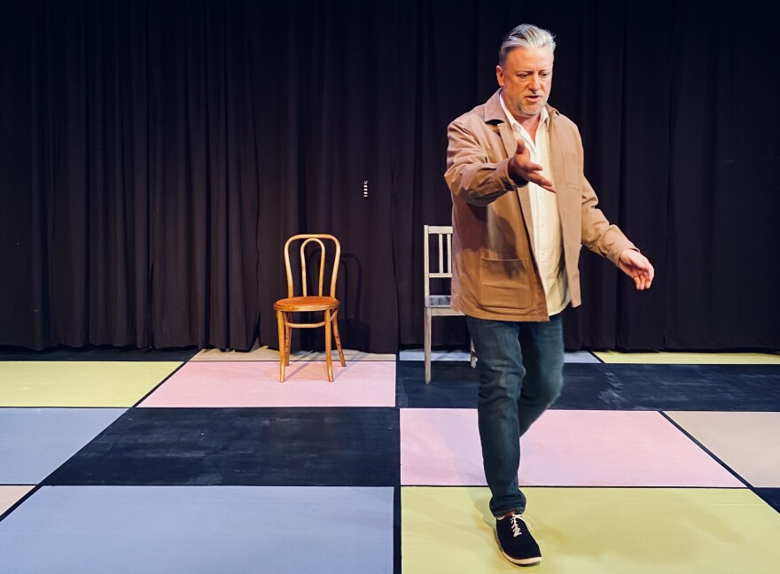 A actor with silver hair and casual clothes gestures to an unseen audience member. The stage is starkly dressed with two chairs and a colorfully painted checkerboard floor.