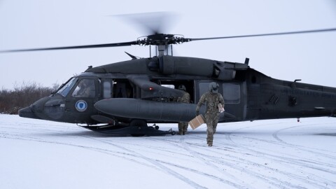 A person wearing camo and carrying a cardboard box walks away from a military helicopter in a snow-covered flat area