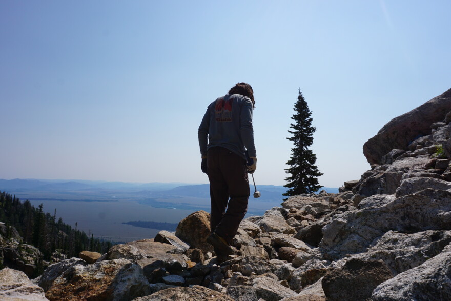 A man stands, looking away with a hammer in his hand, atop rocky boulders, with a view of an open landscape in the background.