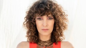 A young woman with curly brown hair looks intensely at the camera with a light background behind her.