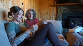  A woman with tattoos on her right arm holds a small baby in her lap on a grey couch. Another woman sits next to her, gazing at the baby.