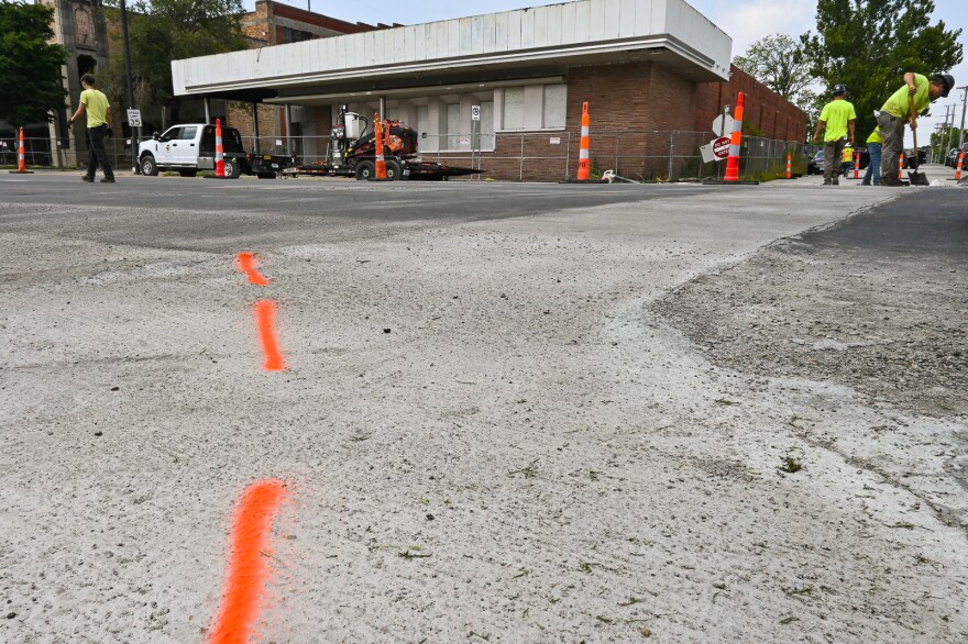 Wide angle view of a street intersection shows people working where a fresh laydown of pavement has been cutaway and patched temporarily.