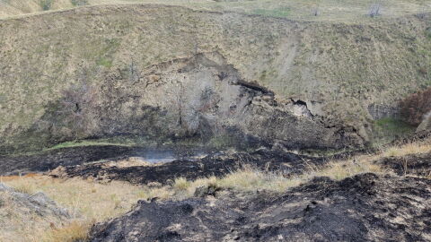 A wildfire caused by an exposed burning coal seam from May of 2025, image depicts blackened and smoking grass near the exposed seam.