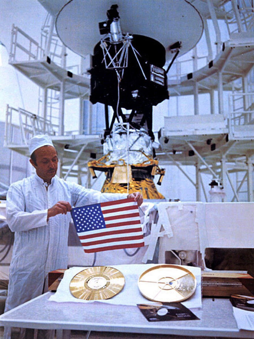 A technician holds a U.S. flag over a gold record containing data from Earth, with a Voyager spacecraft in the background.