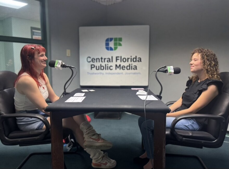 Two young women sit across each other, microphones are in front of them, and they are smiling.