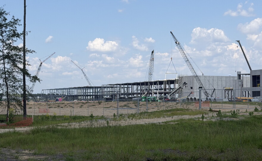 Construction cranes surround two buildings made of steel and concrete. The construction site is surrounded by a chain link fence. 