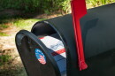 A black mailbox with an "I voted" sticker on it, slightly ajar. There is a mail-in ballot inside and the red mailbox flag is up.