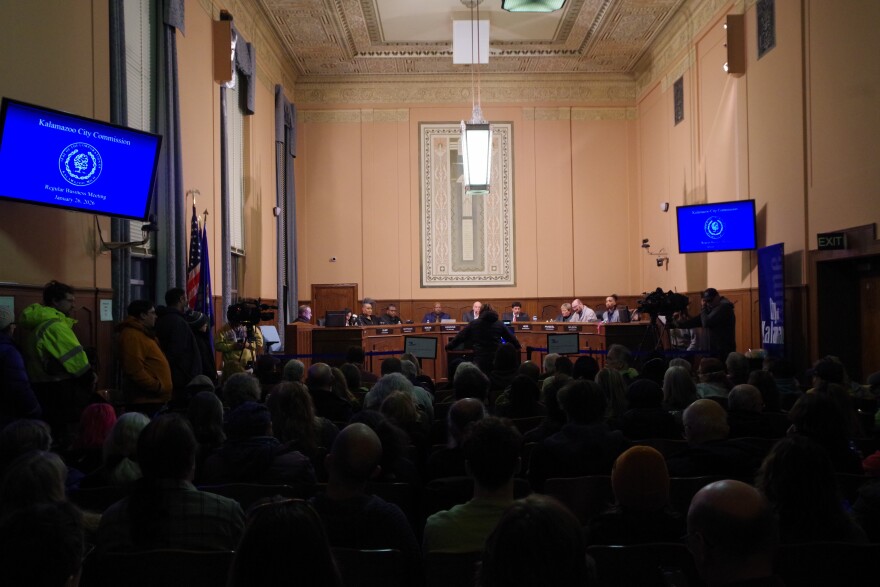 In a large room with an ornate ceiling, commissioners sit at a brightly-lit panel table at the front of the room. In front of them, a crowd of people sitting can be seen in silhouette, as well as a speaker standing at the podium. Several people are lined up against the wall, waiting to speak. 